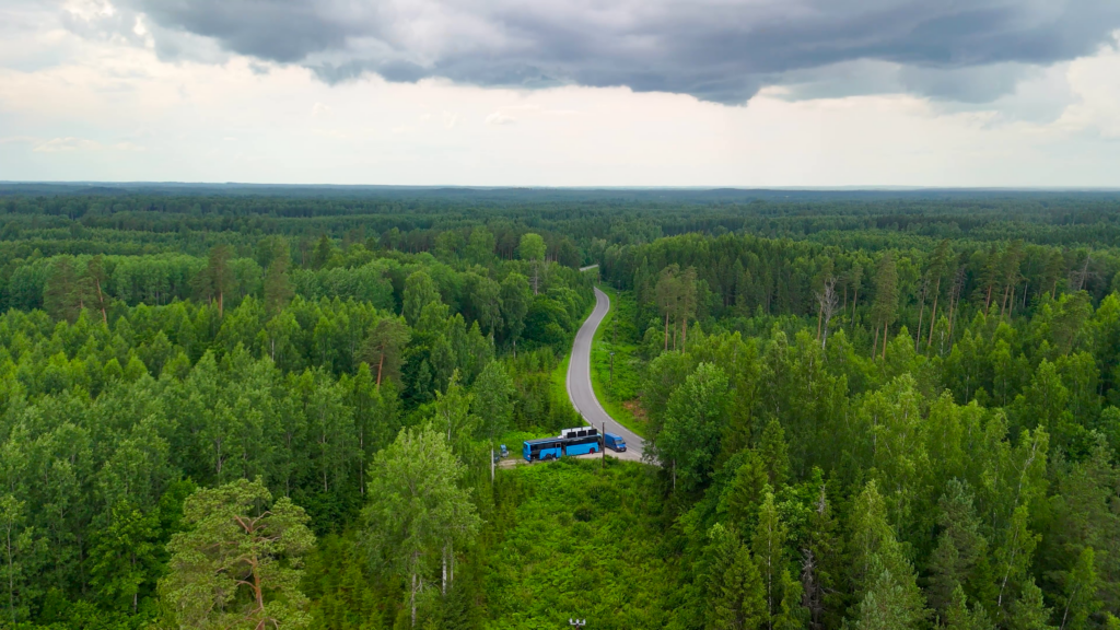 Droonivaade sinisele ülekandebussile ja kaubikule kurvilise metsatee ääres tumedate pilvede all.