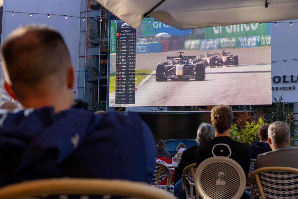 Vaade kohvikus istuvate pealtvaatajate õlgade tagant suurele ekraanile, millel sõidavad F1 autod.